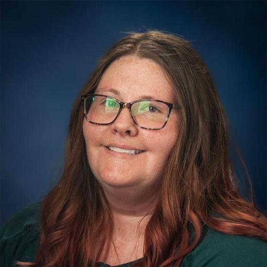 A smiling woman with long brown hair and glasses, wearing a dark green top, is set against a dark blue background.